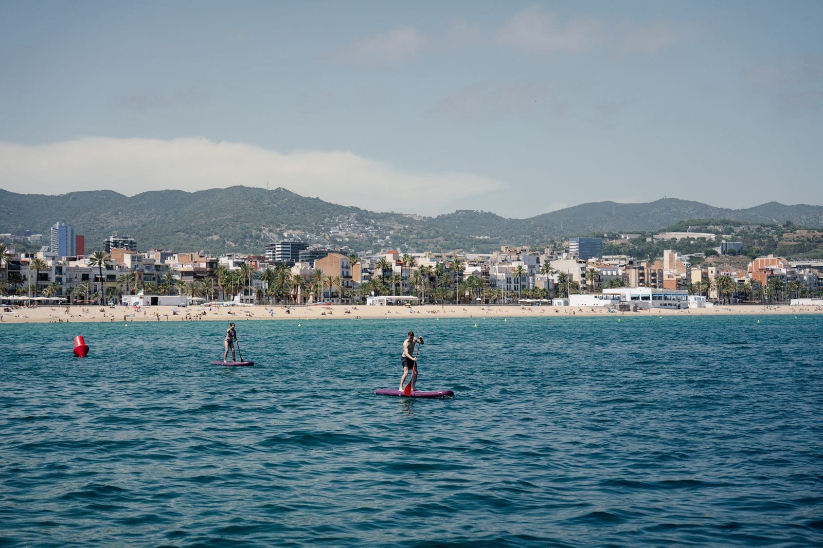 Paddleboarders on calm water beside the GrowBoat catamaran with the Barcelona beachfront and skyline behind